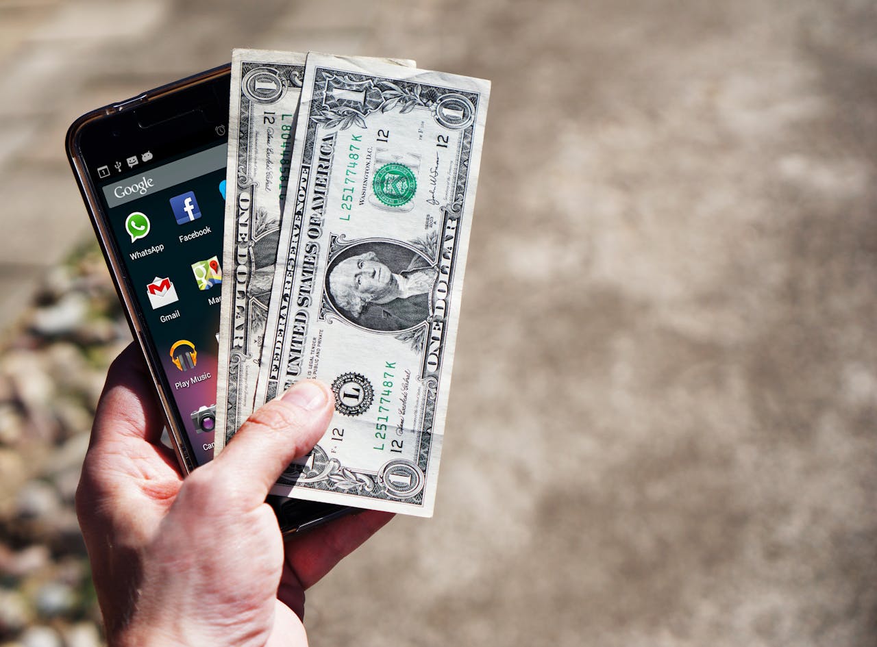 menu-10 Close-up of a hand holding US dollar bills and a smartphone outdoors, showcasing financial technology.