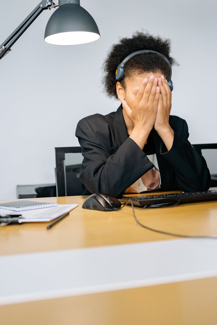 menu-02 A tired call center employee covering her face with hands in frustration, wearing a headset in an office.
